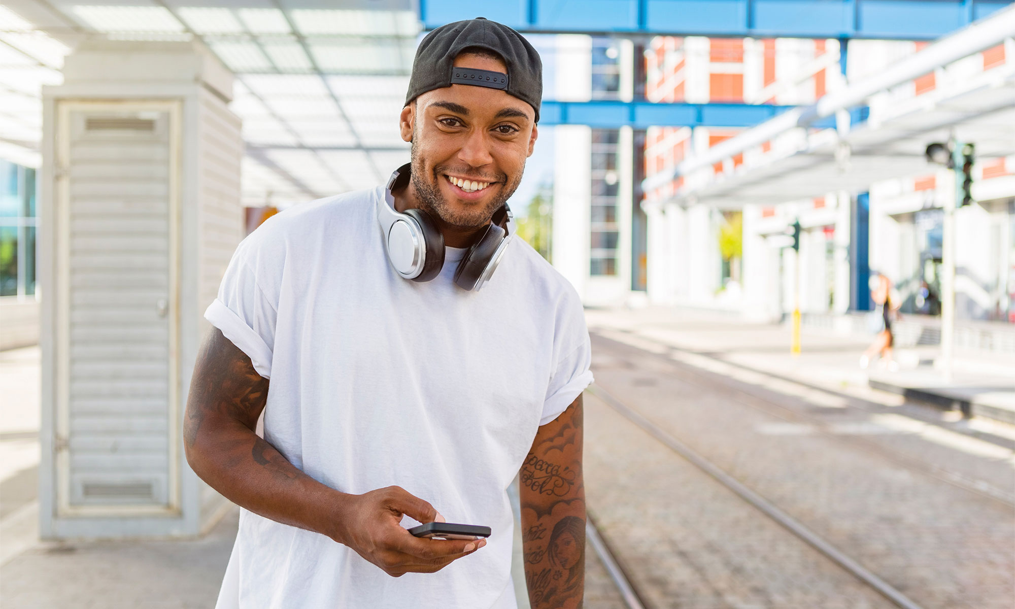 Smiling young man with headphones and smartphone waiting at tram stop.