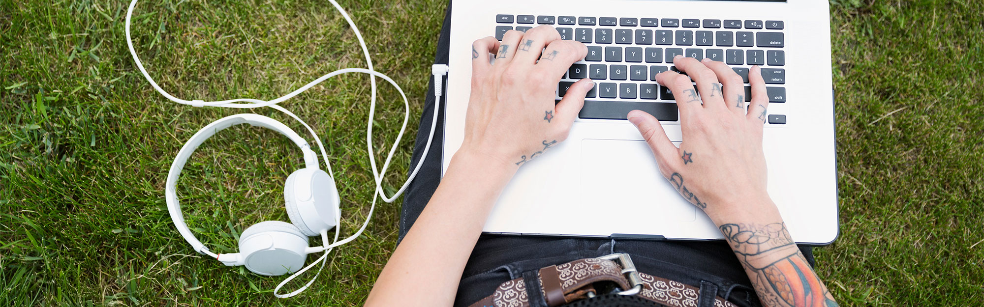 Person med tatueringar sitter i gräset med en laptop i knät